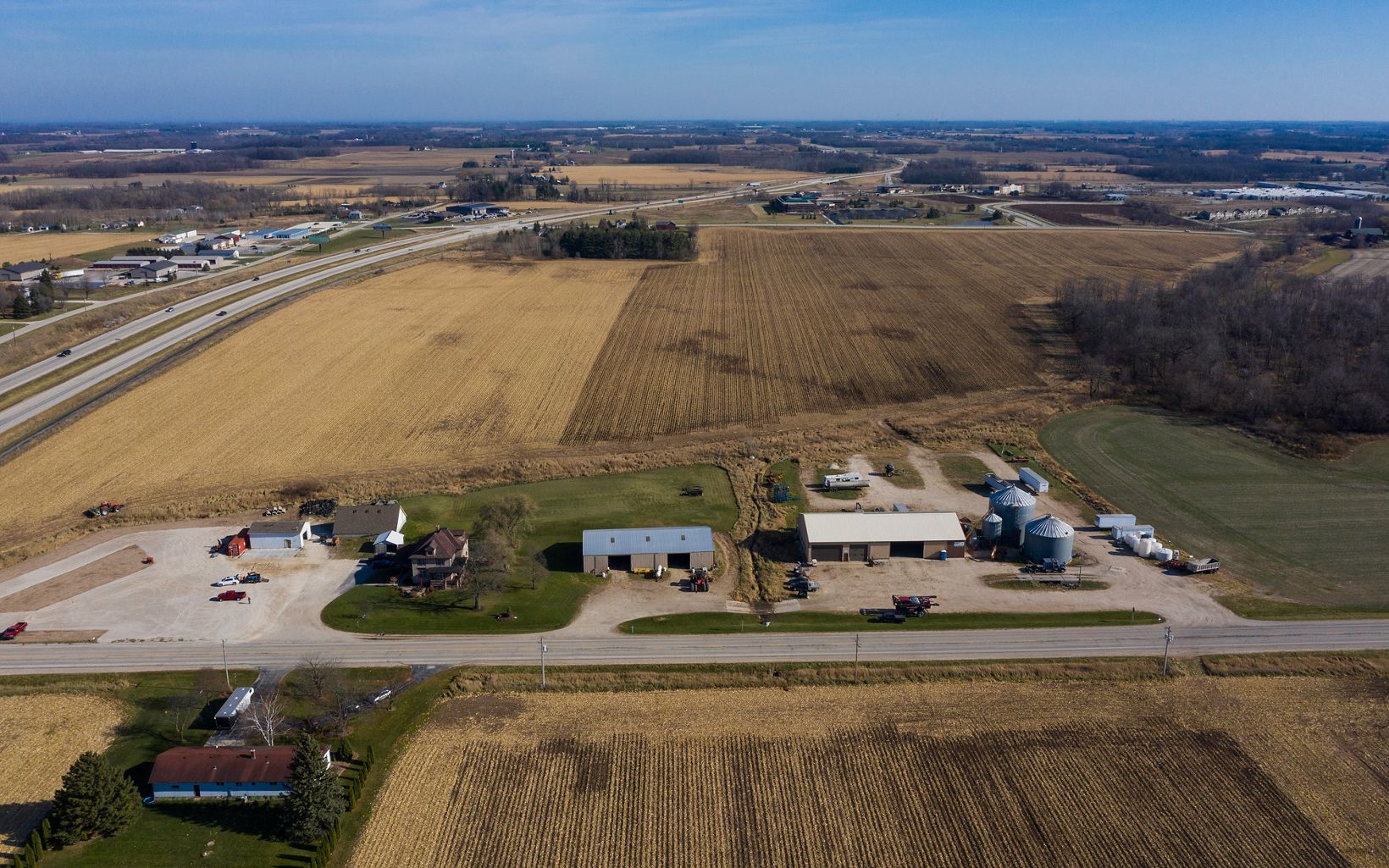Aerial view of several farm buildings and silos, with a road running horizontally in front of them and large golden farm fields behind them.