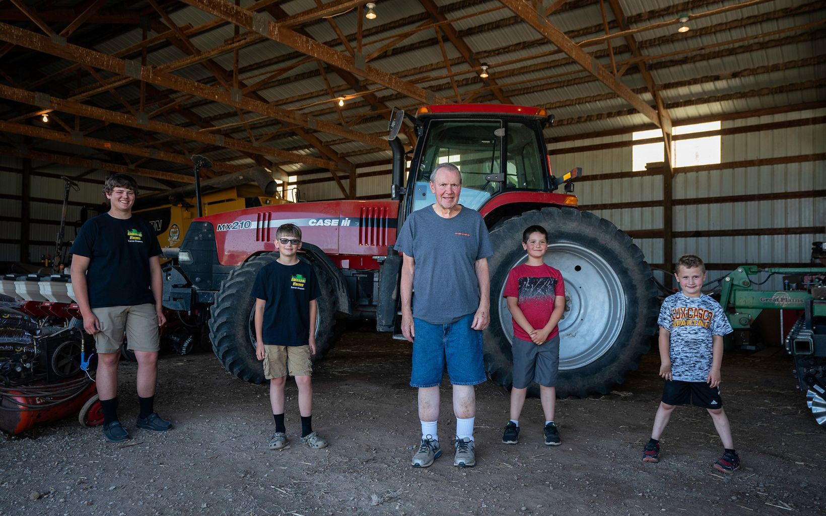 Allen Guilette stands in front of a red tractor in a farm building; he is in the center and two of his grandsons stand on his left, and two stand on his right. 