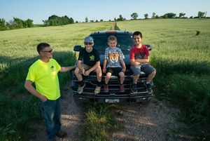 Nick Guilette stands next to a pickup truck on a dirt road in a green field; three young boys sit on the hood of the truck.
