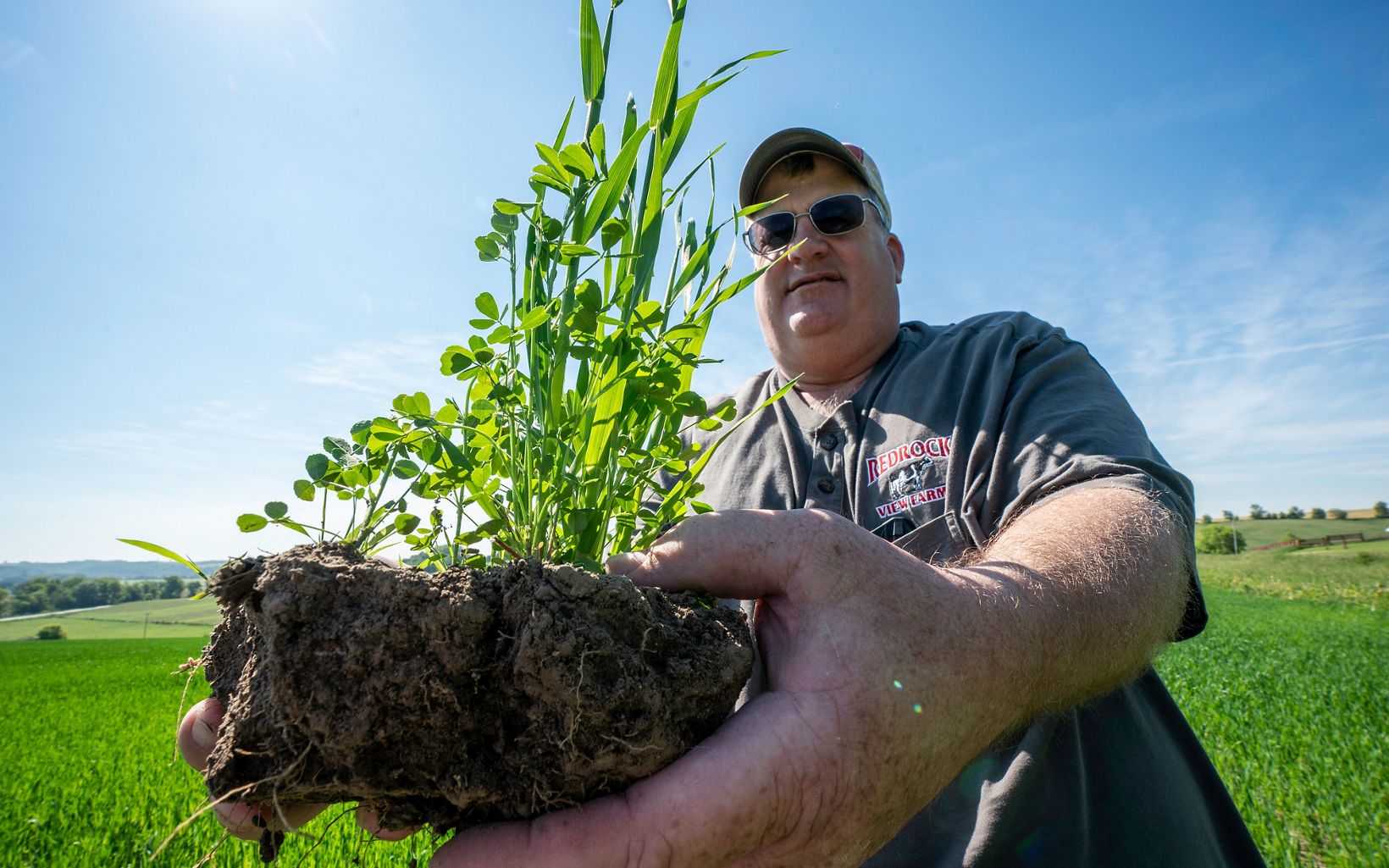 Steve Carpenter stands in a green field and holds up to the camera a large clump of soil with plants growing out of it.