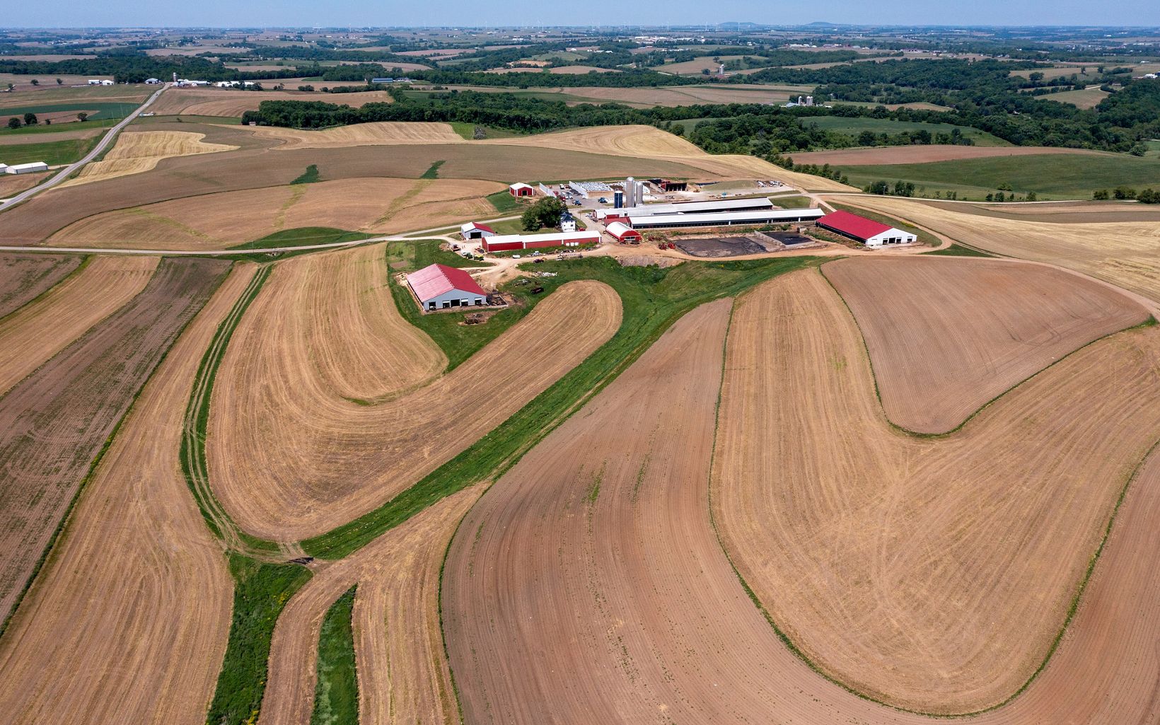 : Aerial view of red-and-white-roofed farm buildings in the middle of broad golden-brown and green farm fields with curvy patterns cut into them.