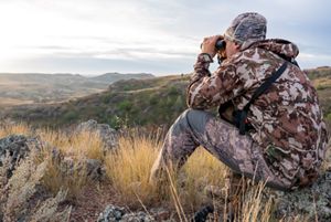 A hunter sits on the rocky ground looking through binoculars.  