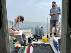 Two men working on a boat. One is standing on the right one is sitting on the left writing in a notebook. 