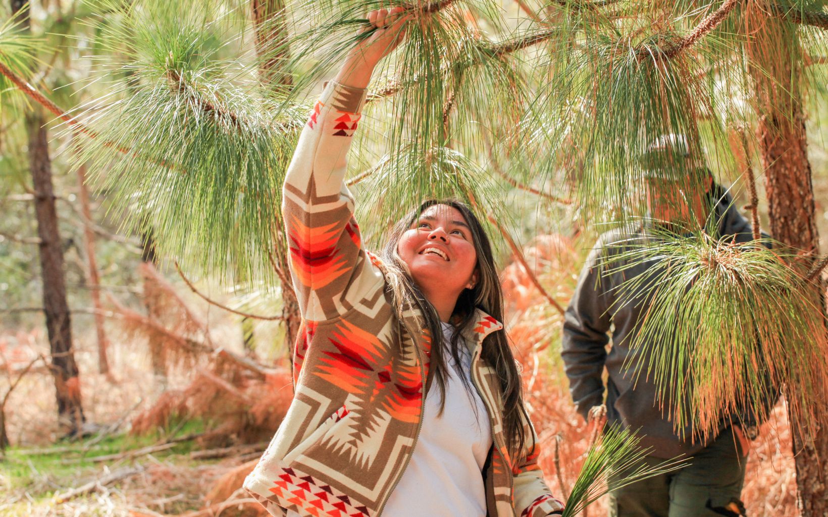 Charity Battise smiles widely as she pulls longleaf needles from a tree with her right hand outstretched. Another harvester stands in the background observing her.