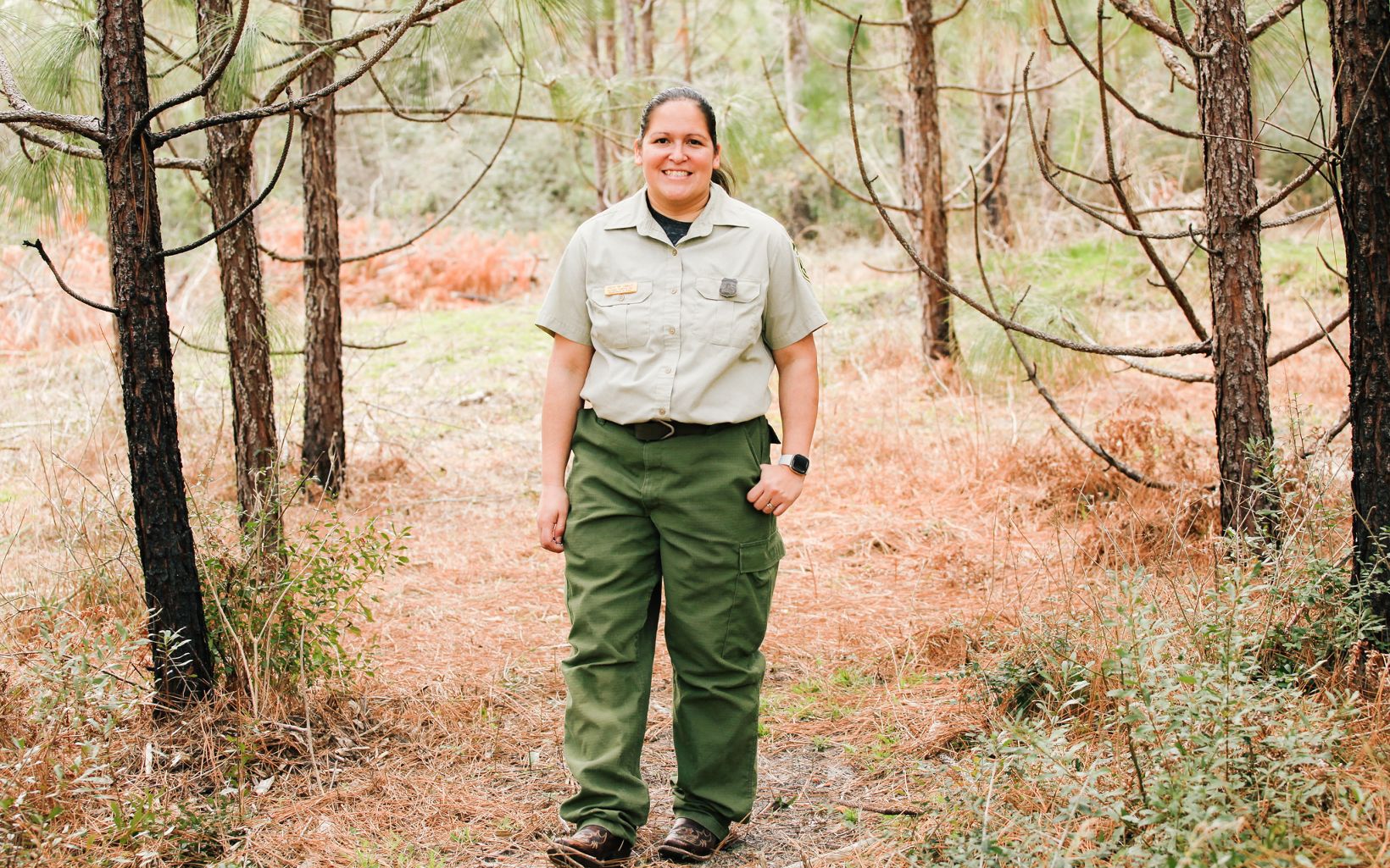 Krista 'KK' Langley, a tribal citizen and USDA Forest Service employee, smiles brightly as she stands among fallen needles and lengthy longleaf trunks.
