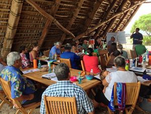 About 20 community members of all ages sitting at tables under a wooden beachside shelter listening to a presentation by a man at the front of the group.