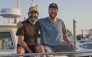 Carl Lobue and Stephen Heck sit on the edge of a boat and smile for the camera.