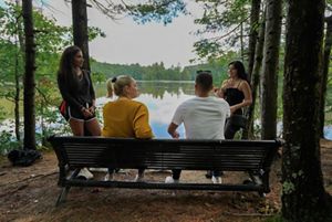 Four young adults gather on a bench in front of a pond.