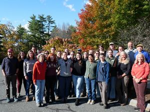 A group of about 30 people standing together smiling outside. Behind them are trees in fall colors.