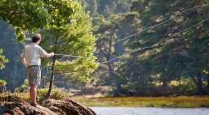 A person stands on a shore and casts a fly-fishing line into a body of water.