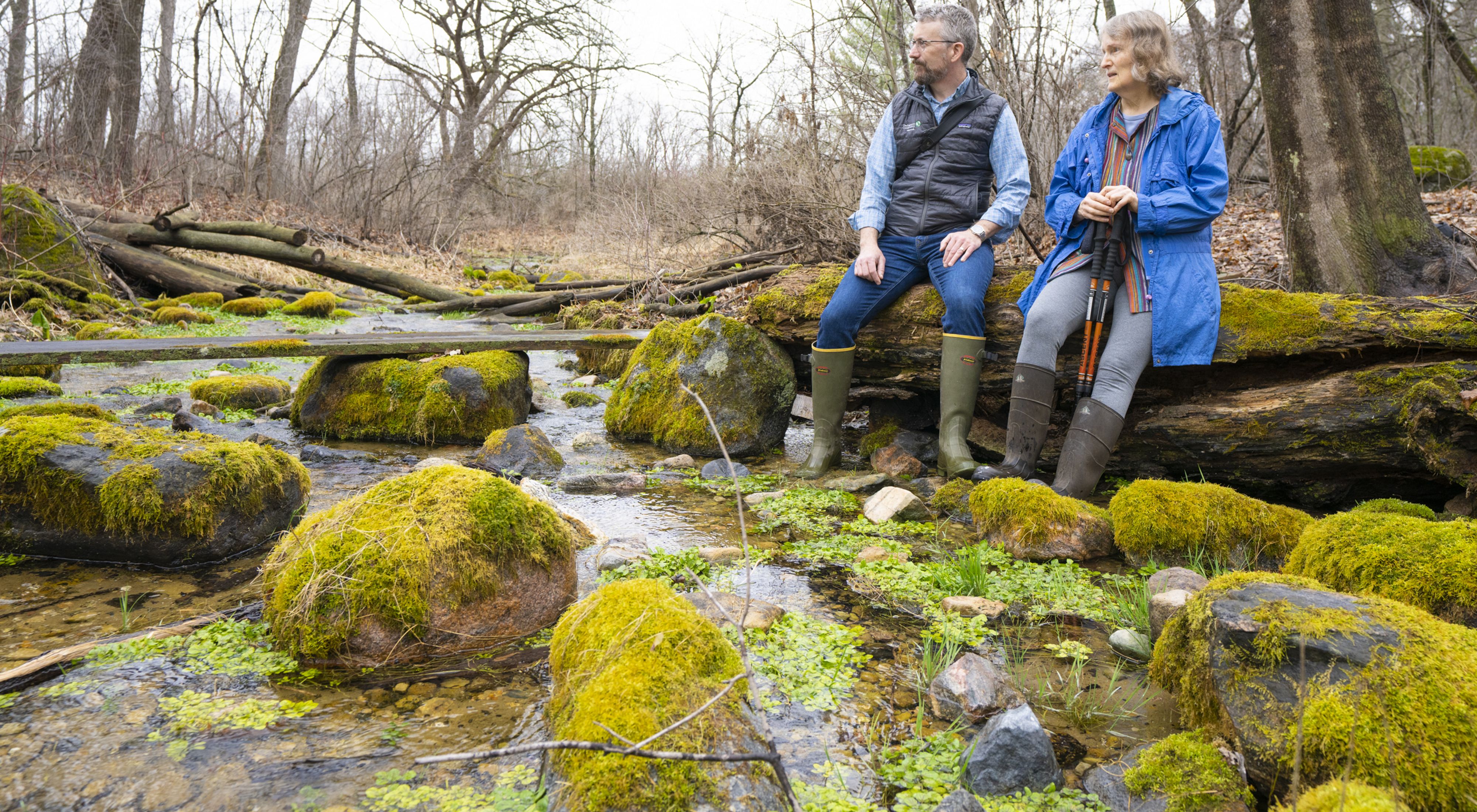 A man and woman in knee-high rubber boots sit on a log with their feet in a shallow, rocky stream.