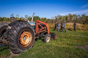 Several people walking together across an open grassy field near an old tractor under a bright blue sky.
