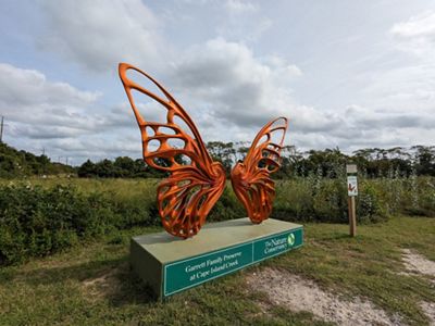 Large orange monarch butterfly statue is situated at the edge of a meadow.
