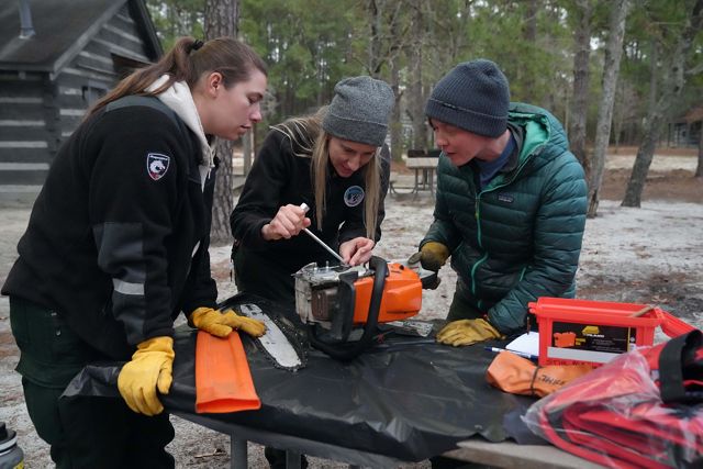 Three women work on a chainsaw over a table outside during a TREX fire training session in North Carolina.