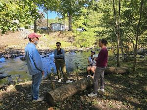 Four people (three stnading, one sitting on a log) gather along the edge of the Mill River in the shade of trees. There are a few buildings in the background on other riverban
