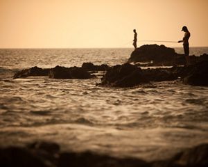 Two people fishing on the rocks of a shoreline at dusk.