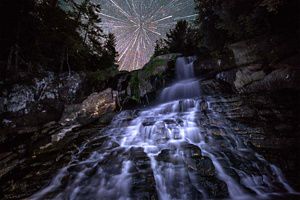 Water cascades down a steep rocky cliff at night, while stars appear to burst overhead like a firework, thanks to detailed photography work.