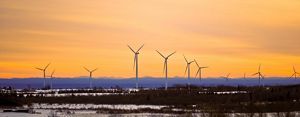 Wind turbines against an orange-hued sky at sunset, standing in a snowy field with ridges of mountains in the distance.