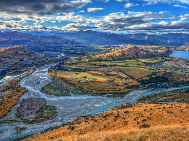 Aerial view of Lake Hayes, New Zealand, showing a meandering river winding around hills with mountain ranges in the distance.
