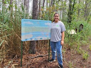 A woman stands next to a sign surrounded by a forest.