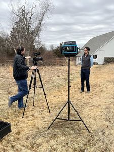 One man talks in front of a video camera while another man stands behind it.