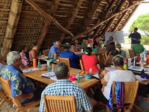 About 20 community members of all ages sitting at tables under a wooden beachside shelter listening to a presentation by a man at the front of the group.