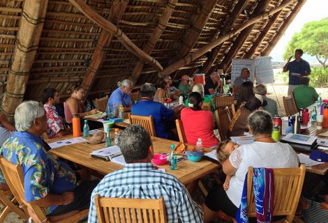 A group of people listen to a presenter at a meeting in an open-air hut.