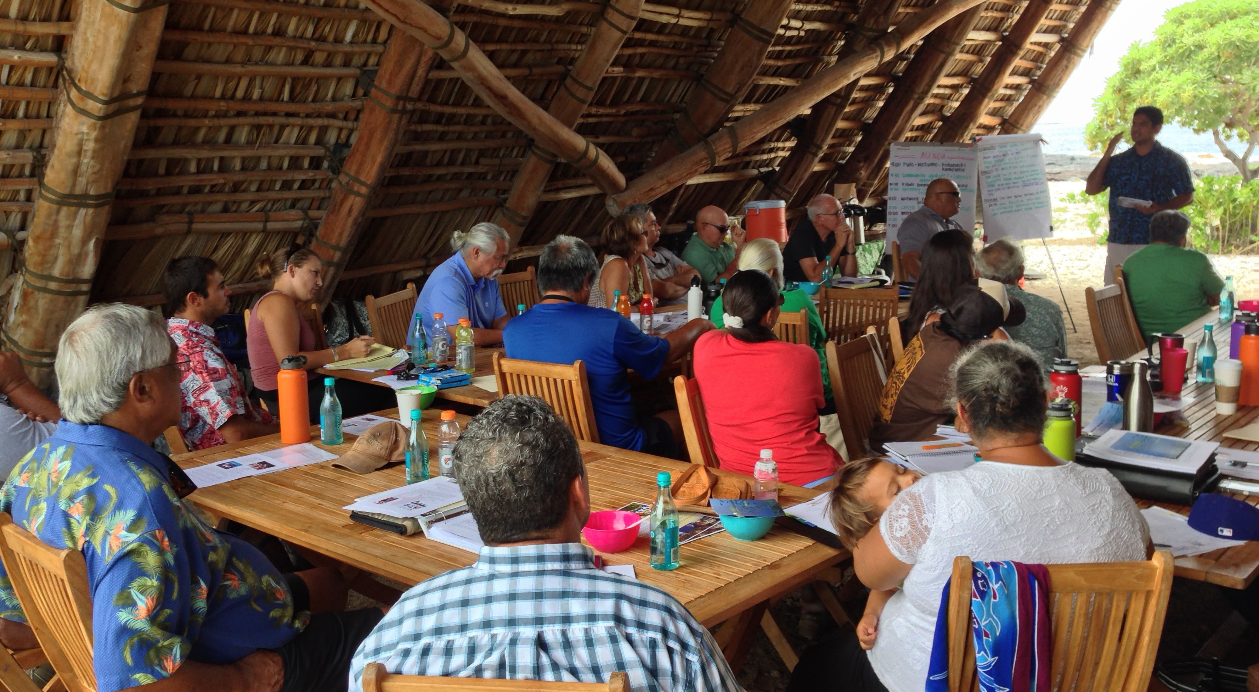 About 20 community members of all ages sitting at tables under a wooden beachside shelter listening to a presentation by a man at the front of the group.