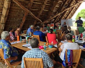 About 20 community members of all ages sitting at tables under a wooden beachside shelter listening to a presentation by a man at the front of the group.