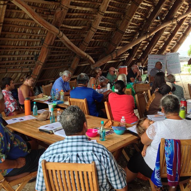 A group of people listen to a presenter at a meeting in an open-air hut.