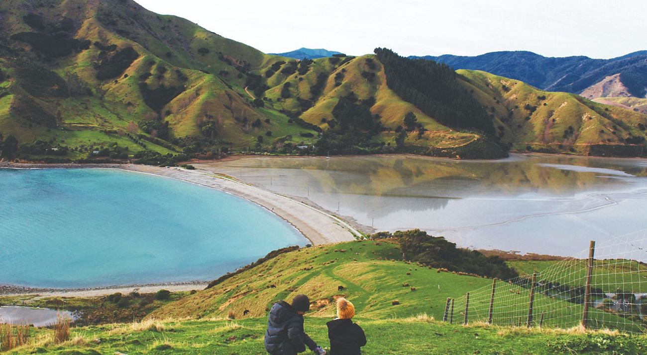 Two people sit at the edge of a wide body of water with mountains in the distance, enjoying the scenic views in Nelson, South Island.