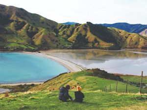 Two people sit at the edge of a wide body of water with mountains in the distance, enjoying the scenic views in Nelson, South Island. 