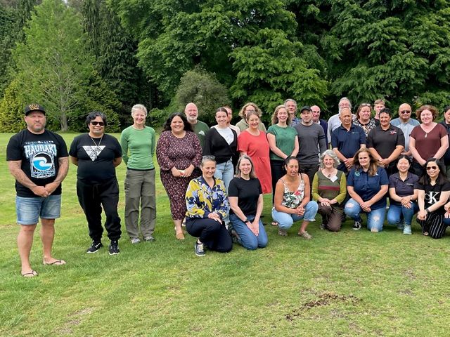 A large group of people stand together for a group photo in a grassy field with a forest behind them.
