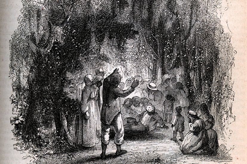 A historical print of an enslaved person's funeral taking place in a heavily wooded grove. Numerous mourners gather around a preacher with raised, outstreched arms. Mourners kneel, pray, and weep. 