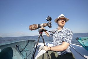 A young man in a blue plaid shirt and white brimmed hat sitting in a boat at sea next to a large camera mounted on a tripod.