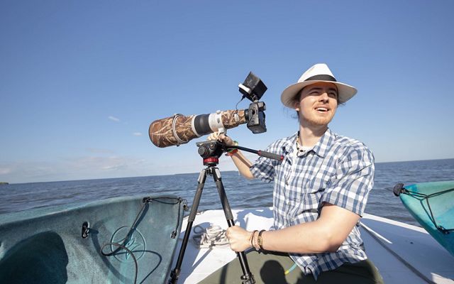 A young man in a blue plaid shirt and white brimmed hat sitting in a boat at sea next to a large camera mounted on a tripod.