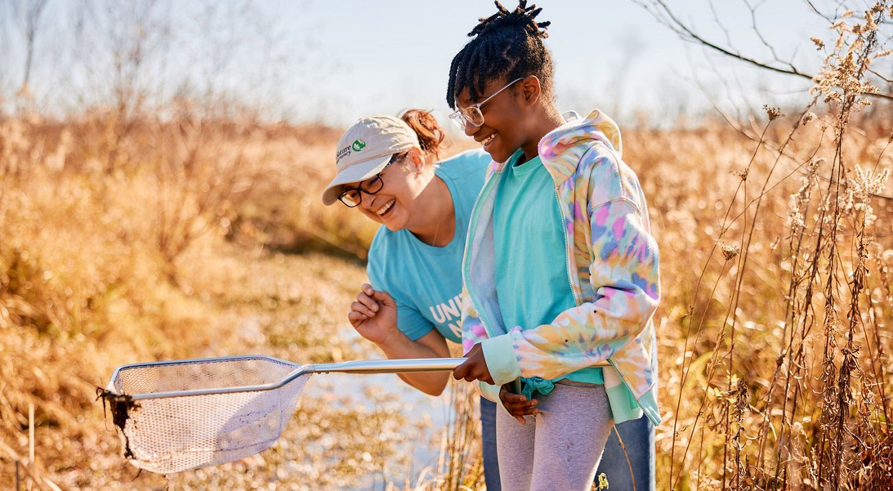 A woman wearing a TNC hat and a young girl smile while observing the contents of a dip net in a field.