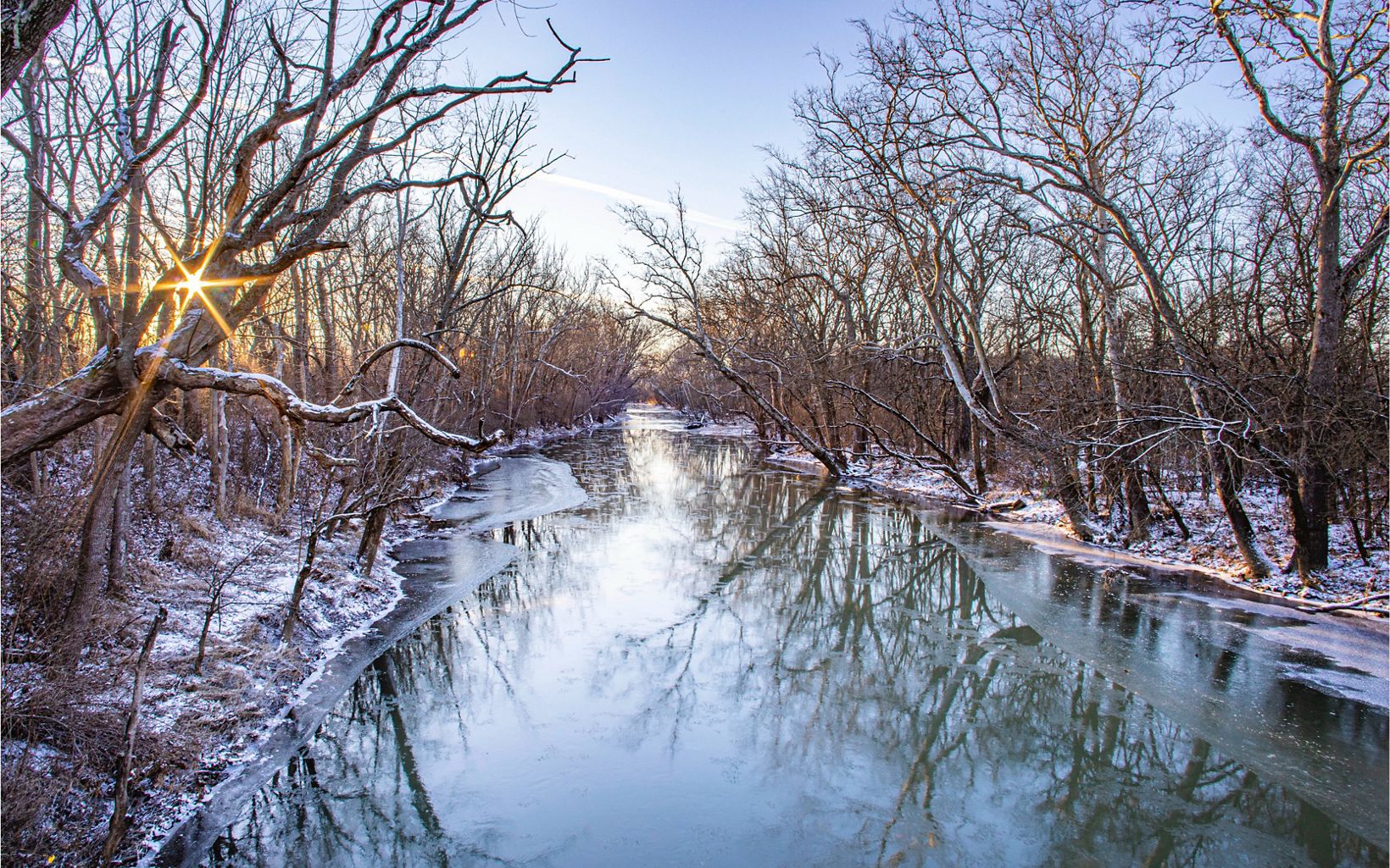 Snow-covered trees along a partially frozen creek at sunset.