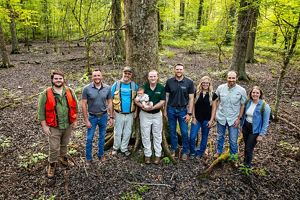 Eight smiling adults stand in forested landscape. Man in center holds infant.