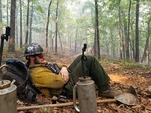 Man sits under a tree next to a drip torch during a controlled burn.