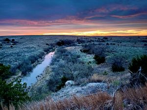 The sun sets behind a calm, narrow creek winding through a vast landscape.