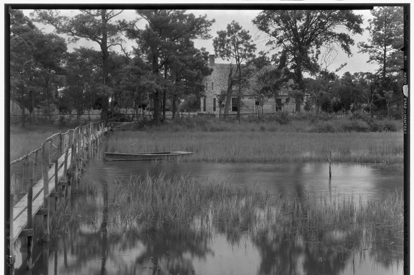 Vintage black and white image of an abandoned single story house nestled in the trees at the edge of a wide creek. 