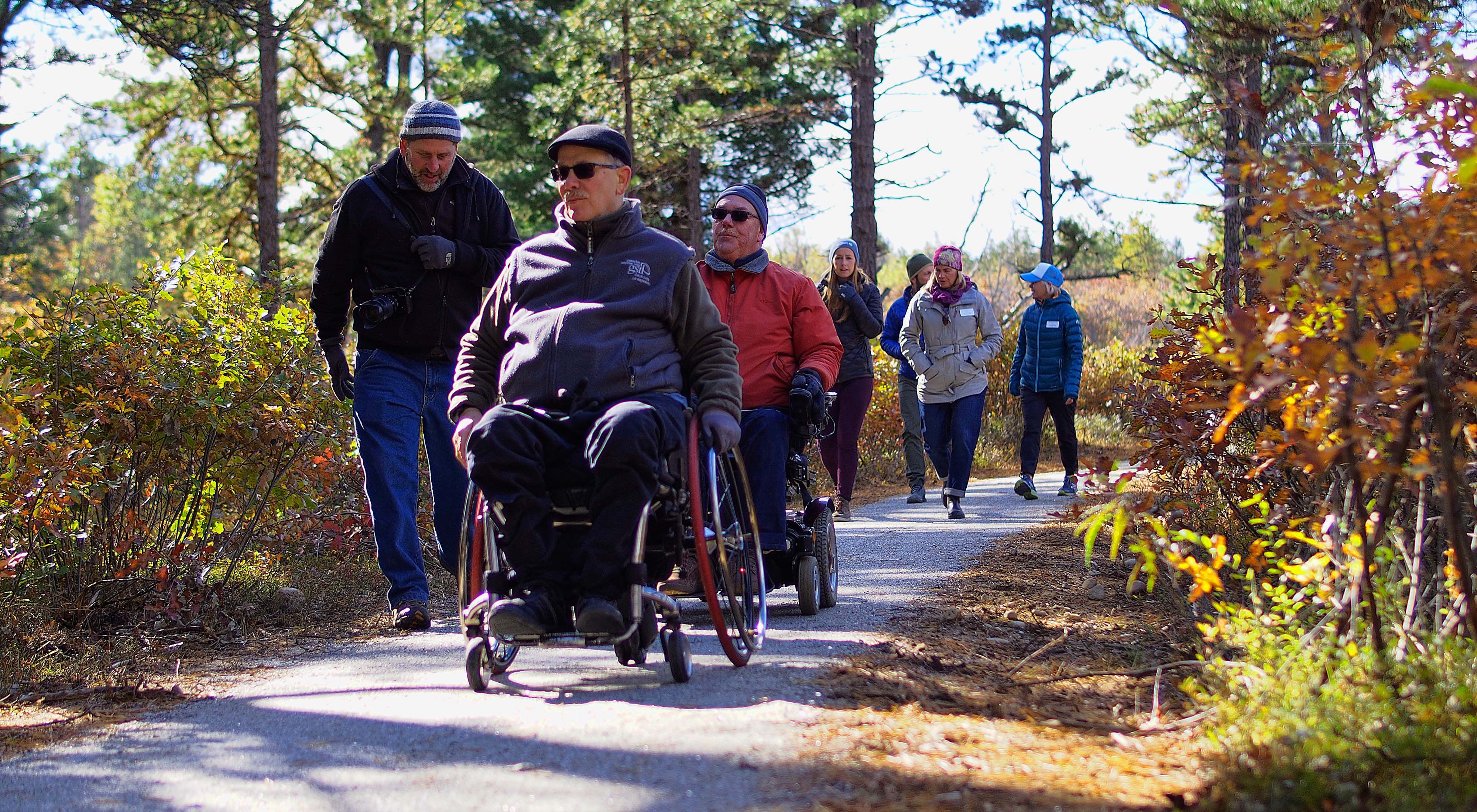 A group including a man using a wheelchair make their way down a path in the woods.