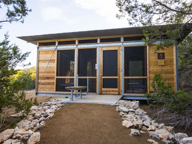 A gravel trail leads to a wooden cabin with a metal roof surrounded by dense brush.