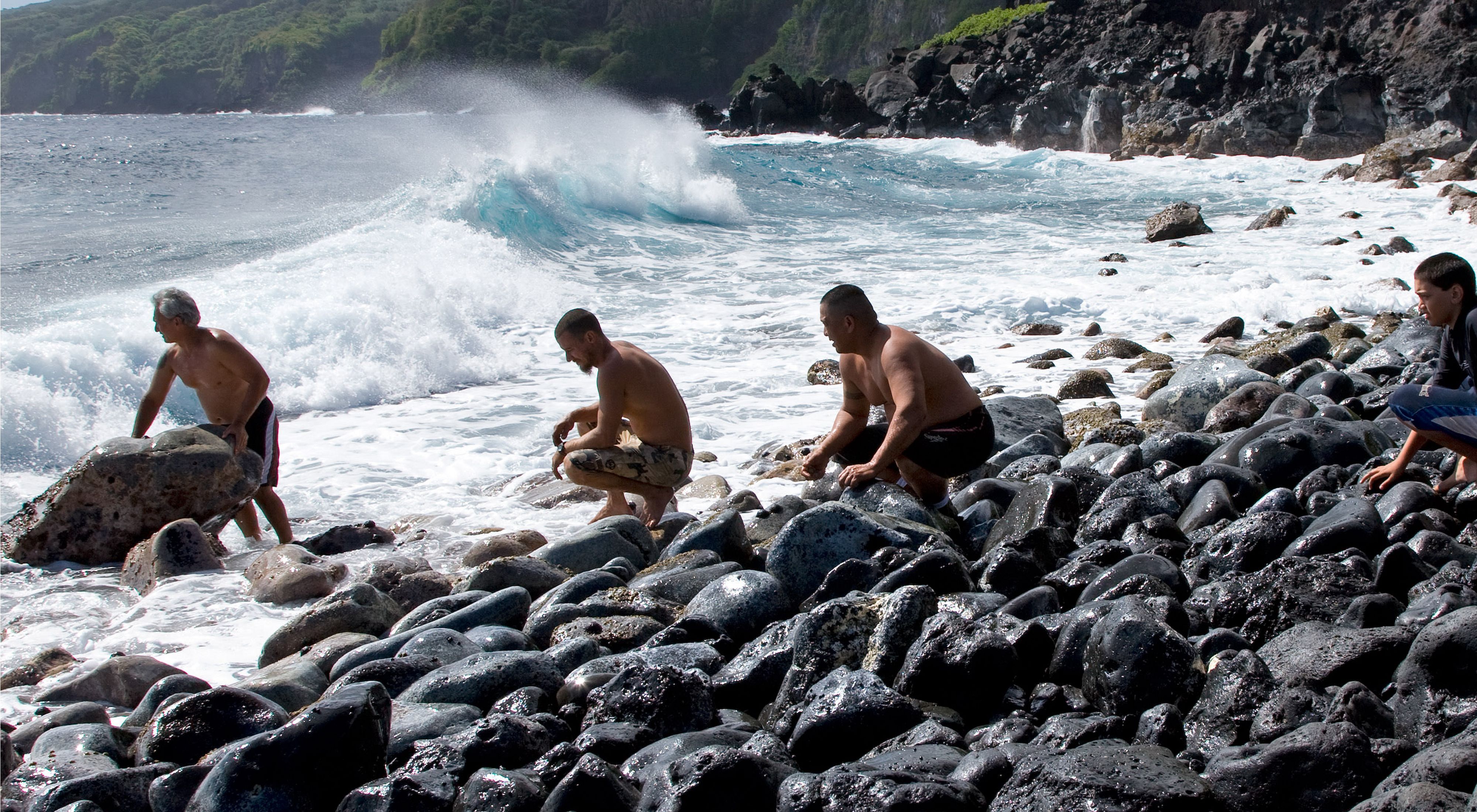 A group of four men crouch together on a rocky ocean coast as the surf crashes around them.