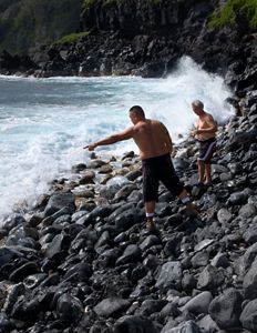 A group of four men crouch together on a rocky ocean coast as the surf crashes around them.
