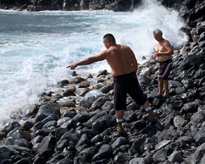 A group of four men crouch together on a rocky ocean coast as the surf crashes around them.