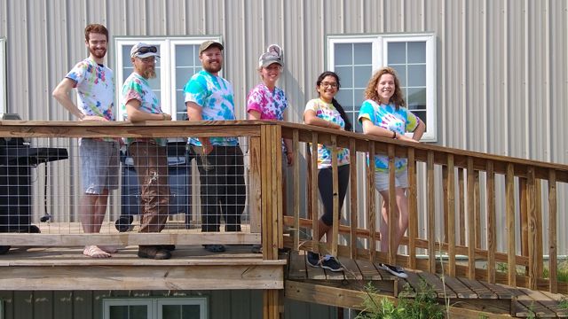 Volunteers in tie-dye shirts pose outdoors on a wooden ramp in front of a building.