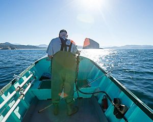 A man wearing waders stands at the bow of a small boat on a wide body of water.