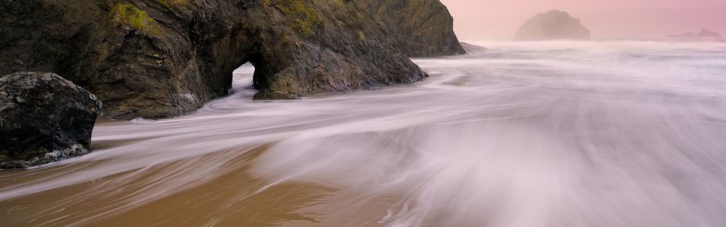 A time-lapse photo of waves coming in at a sandy beach bordered by rugged boulders and cliffs.